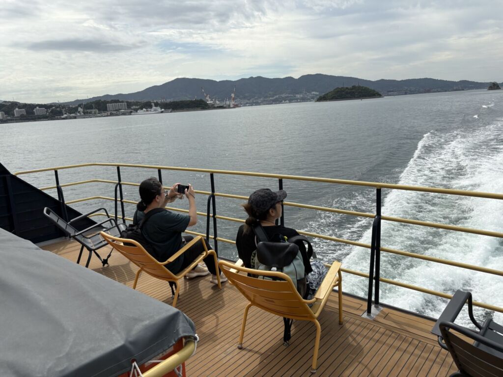 SEA SPICAのデッキから望む瀬戸内海。黄色い手すりの向こうに白い航跡が伸び、対岸に呉の街と山並みが見える