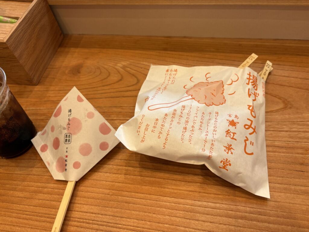 紅葉堂の揚げもみじ。イートインスペースの木のテーブルに、串に刺さった揚げもみじの包みとアイスコーヒーが置かれている